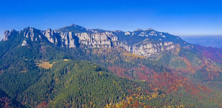 Aerial view of rocky mountain and colored forest in Romanian Carpathians, beautiful autumn landscape with blue sky.