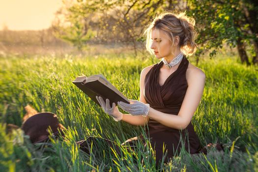 young blonde girl in a brown vintage dress and top hat reads a book in spring garden