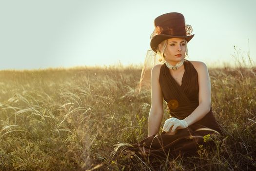 young blonde girl in a brown vintage dress and top hat in a feather grass