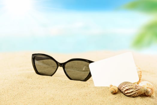 Vacation concept image - sunglasses, shells and blank white card on a sandy tropical beach in close-up and on a blurred coastline background.