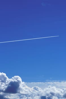 The trail of a flying airplane in a clear blue sky over large clouds.