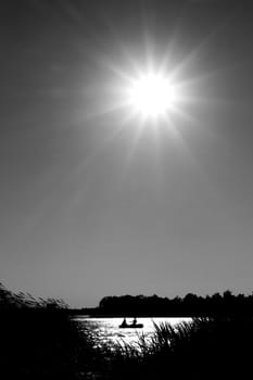 Silhouettes of two people on a rubber boat in a sunny reflection & cane foreground on the surface of a lake. The bright sun in the sky. Black & White.