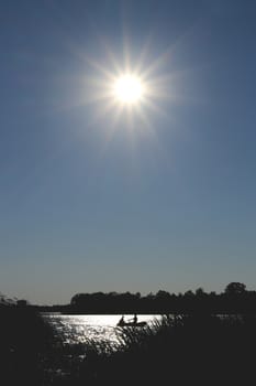 Silhouettes of two people on a rubber boat in a sunny reflection & cane foreground on the surface of a lake. The bright sun in the sky.