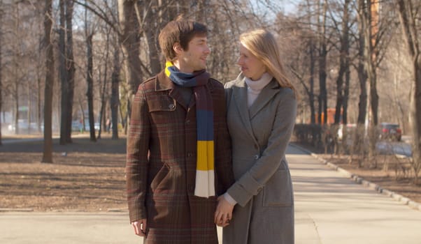 Beautiful couple in love walking in the park, young blond girl in a gray coat and man in brown coat and colorful scarf holding hands, smiling and rejoicing early spring sunny day.