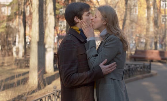 Beautiful couple in love kissing in the park, young blond girl in a gray coat and man in brown coat and colorful scarf. Spring sunny day.