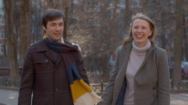 Beautiful couple in love walking in the park, young blond girl in a gray coat and man in brown coat and colorful scarf holding hands, smiling and rejoicing early spring sunny day.