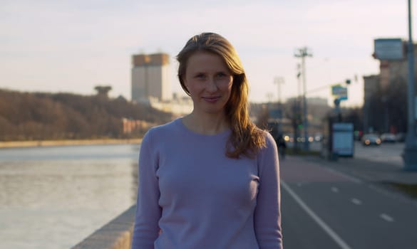 Portrait of a young smiling beautiful woman standing on the embankment near the river against the background of the city. Blond woman with long hair outdoors