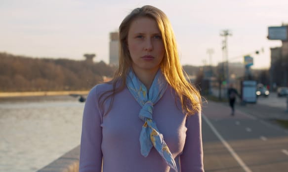 Portrait of a young smiling beautiful woman standing on the embankment near the river against the background of the city. Blond woman with long hair outdoors