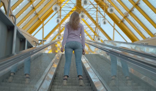 A young woman standing on the moving escalator. Rear view, follow shot. Blond woman with long hair inside modern building