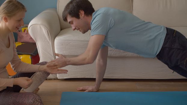 A young cheerful family spends time at home. The husband does push-ups with clapping, the wife sitting nearby. Both laughing. Coronavirus epidemic