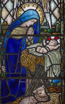 Historic stained glass window showing the Virgin Mary praying over the baby Jesus lying in a straw filled manger with lambs.