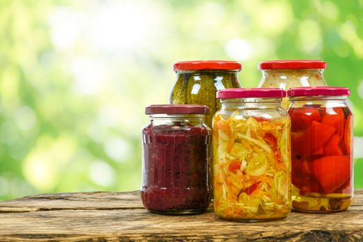 Composite image of jars with variety of homemade pickled vegetables on a wooden vintage table and on the blurred green garden background.