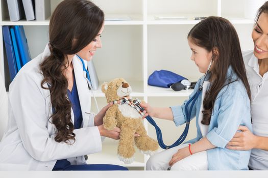 Cute small child patient pretending to be doctor, holding teddy bear toy at meeting with general practitioner. Friendly nurse showing how stethoscope works to smiling little girl at checkup in clinic.