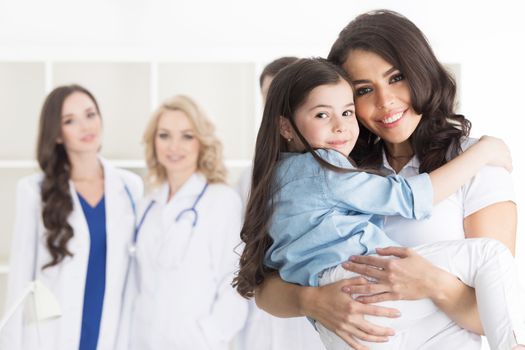 Mother and daughter in medical clinic, team of doctors on background