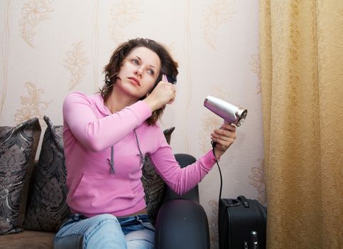 young brunette woman in pink blouse and blue jeans dries the hair dryer sitting on the couch