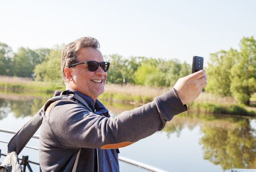 elderly man makes selfie with mobile phone near the river on summer day