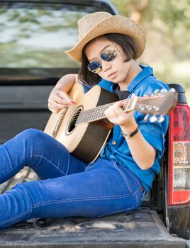 Young asian women short hair wear hat and sunglasses playing guitar ,sit on pickup truck in countryside Thailand