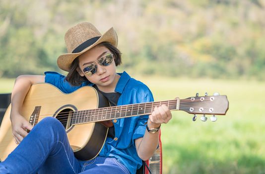 Young asian women short hair wear hat and sunglasses playing guitar ,sit on pickup truck in countryside Thailand