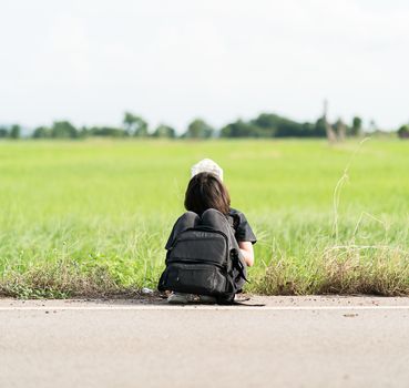 Young asian woman short hair and wearing sunglasses sit with backpack hitchhiking along a road wait for help in countryside Thailand