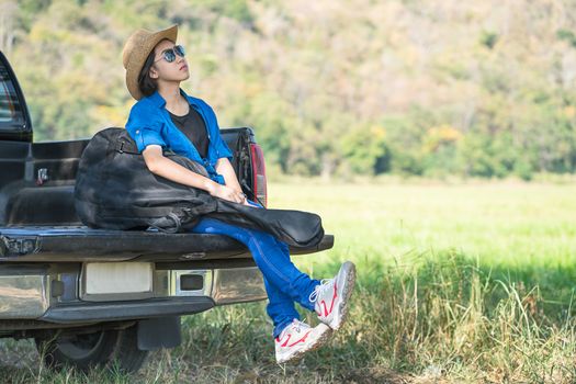 Young asian women short hair wear hat and sunglasses carry her guitar bag ,sit on pickup truck in countryside Thailand