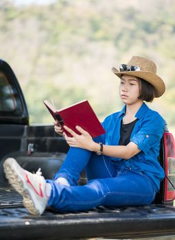 Young asian women short hair wear hat and sunglasses read a book ,sit on pickup truck in countryside Thailand