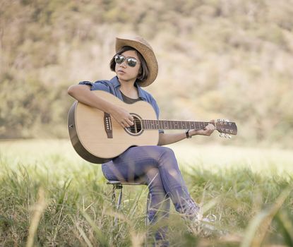 Young asian women short hair wear hat and sunglasses sit playing guitar in grass field countryside Thailand