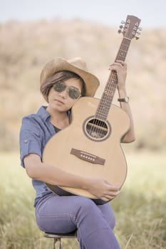 Young asian women short hair wear hat and sunglasses sit playing guitar in grass field countryside Thailand