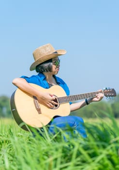 Young asian women short hair wear hat and sunglasses sit playing guitar in grass field countryside Thailand