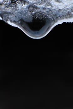 Cold clear rain drop with ice reflection inside. Handing down water drop from ice top on black background. Macro closeup.
