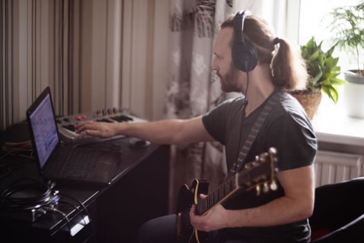 Bearded man musician playing music and composing a song with electric guitar piano and laptop computer while sitting in living room, corona virus quarantine stay home concept