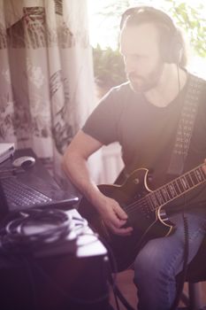 Bearded man musician playing music and composing a song with electric guitar piano and laptop computer while sitting in living room, corona virus quarantine stay home concept