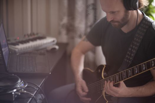 Bearded man musician playing music and composing a song with electric guitar piano and laptop computer while sitting in living room, corona virus quarantine stay home concept