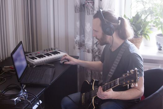 Bearded man musician playing music and composing a song with electric guitar piano and laptop computer while sitting in living room, corona virus quarantine stay home concept