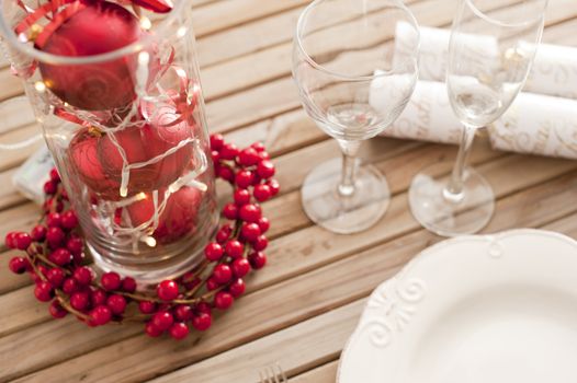 Decorative red and white themed Christmas table setting with an empty clean plate, wineglass and baubles in a glass container surrounded by berries, high angle view
