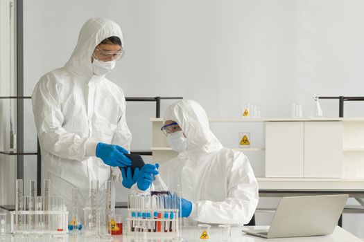 Epidemiological researchers in virus protective clothing use tablet computers to research chemical compounds on the internet. Working atmosphere in chemical laboratory.