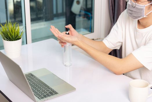 Asian business young handsome man glasses working from home office he quarantines disease coronavirus or COVID-19 wearing protective mask and cleaning hands with sanitizer gel on front laptop computer