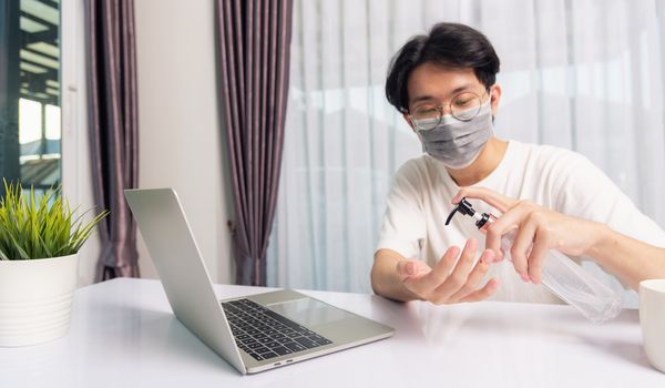 Asian business young handsome man glasses working from home office he quarantines disease coronavirus or COVID-19 wearing protective mask and cleaning hands with sanitizer gel on front laptop computer