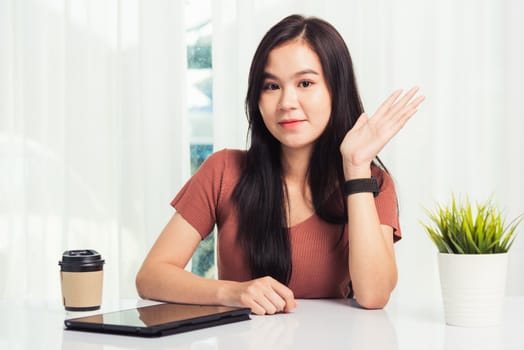 Work from home, Asian business young beautiful woman smiling sitting on desk workspace raise hand say hello communicating with team video chat or conferencing on digital tablet computer at home office