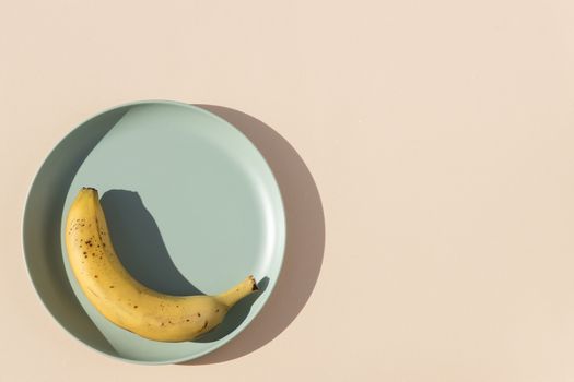Minimalist image of a banana on a light green plate with a beige background and hard shadows of daylight. Top view.
