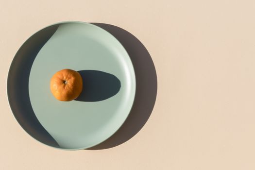 Minimalist image of a mandarin on a light green plate with a beige background and hard shadows of daylight. Top view.
