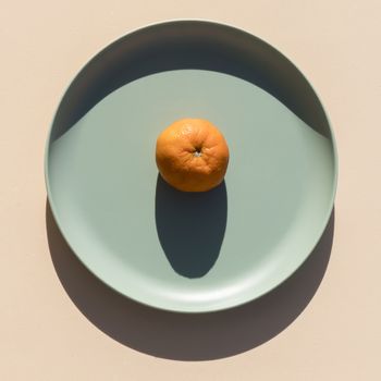 Minimalist image of a mandarin on a light green plate with a beige background, with hard shadows of daylight. Top view.