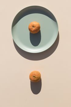 Minimalist and symmetrical image of two mandarins, one on a light green plate and the other on a beige background, with hard shadows of daylight. Top view.