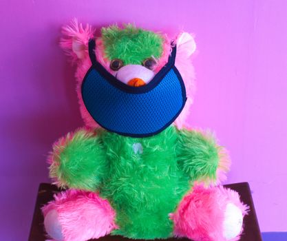 A pink and green colored teddy bear wearing a mask in mouth indicating to be safe from corona virus