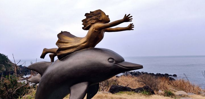 A girl statue with outstretched arms looking at the blue sky on a dolphin structure in Jeju Island, South Korea