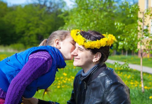 daughter kisses mom on spring day in the park. her mother wore on her head a crown of dandelions