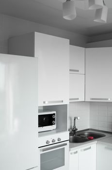 Comfortable white kitchen with a white lacquered facades. Modern kitchen clean interior design. Refrigerator, kitchen oven, microwave oven and sink. Kitchen supplies