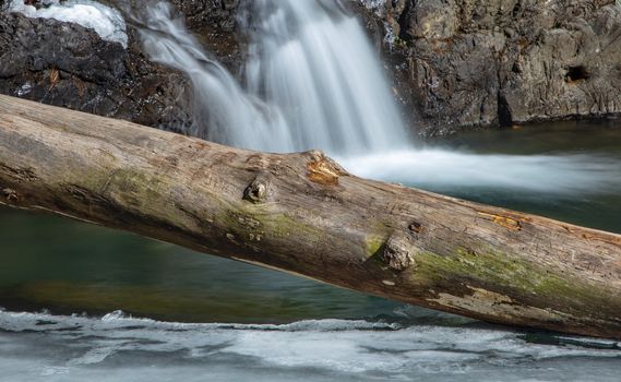A wooden log lays on a thin sheet of ice on top of a river in Arkansas with a waterfall in the background.