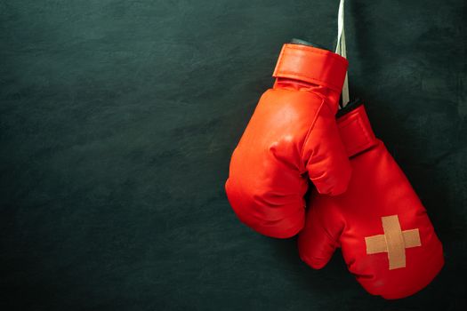 Red boxing gloves hung on black cement wall in darkness with lighting. Adhesive plaster across each other on boxing gloves. Idea of hurt or combat losing business rivals. Fighting giving up boxing.