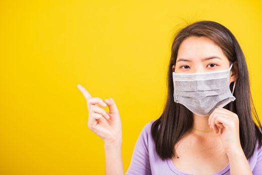 Asian young teen woman wearing face mask protective against coronavirus or COVID-19 virus or filter dust, air pollution her point finger to side space, studio shot isolated yellow background