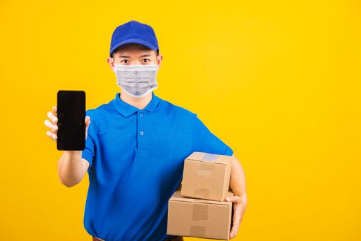 Asian young delivery worker man t-shirt and cap uniform wearing face mask protective show front black screen of smart mobile phone under coronavirus or COVID-19, studio shot isolated yellow background